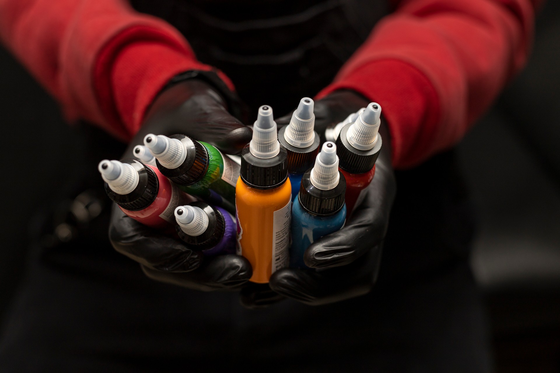 Male tattoo artist holding bottles of tattoo ink of various colors. Selective focus on the jars, black background, wearing red sweater with black gloves. Body art concept